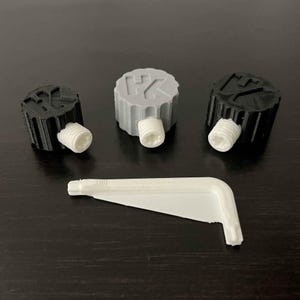May include: Three 3D-printed items: two black and one gray cylindrical caps with white screws, and a white angled tool. The caps have textured tops with symbols. The tool is white and has a right-angle shape.
