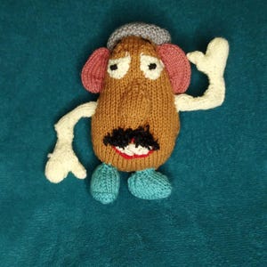 Puede incluir: Un juguete de punto de Mr. Potato Head, un personaje popular de la franquicia Toy Story. El juguete es marrón con un sombrero gris, orejas rosas, un bigote negro y zapatos azules.