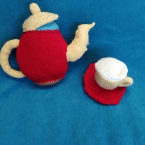 May include: A knitted red teapot with a cream lid and a blue stripe, and a knitted cream teacup with a white rim and a red saucer.