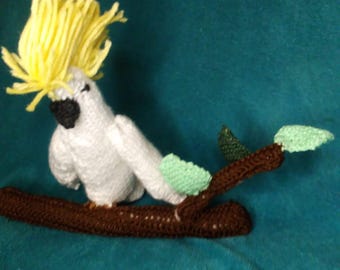 May include: A crocheted white cockatoo with a yellow crest perched on a brown branch with green leaves.