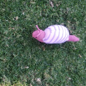 May include: A handmade, knitted snail toy with a pink head and tail, and a white and lavender striped shell. The snail is resting on a bed of green grass.