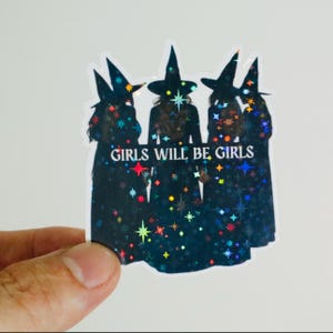 May include: A sticker featuring three figures in witch hats and robes, with a starry, holographic effect. The text "GIRLS WILL BE GIRLS" is printed in white across the center of the sticker. The sticker has a white border.