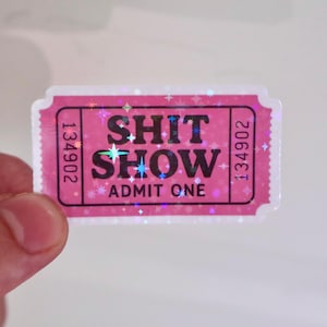 Op de afbeelding: Een roze ticketvormige sticker met de woorden "SHIT SHOW" in vette zwarte letters en "ADMIT ONE" eronder. Het ticket heeft een holografisch effect en de nummers "134902" zijn aan weerszijden gedrukt.