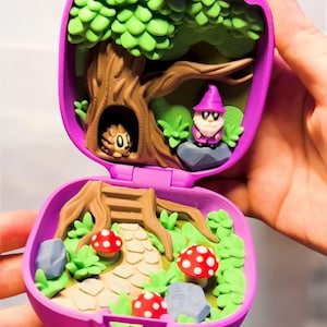 May include: A purple, hinged miniature playset featuring a forest scene. Inside, a tree with a hedgehog in its hollow and a gnome on a rock. The lower half shows a path, mushrooms, and greenery. The playset is held open.