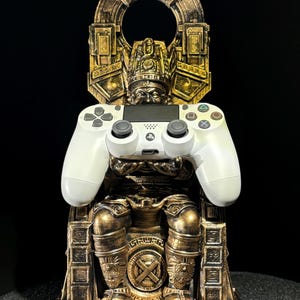 May include: A gold-colored statue of a figure sitting on a throne, holding a white PlayStation 4 controller. The statue is detailed with intricate carvings and a crown on the figure's head.