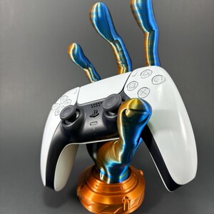 May include: A white PlayStation 5 controller is held by a 3D printed hand stand. The hand is made of blue and orange filament and has a copper colored base.