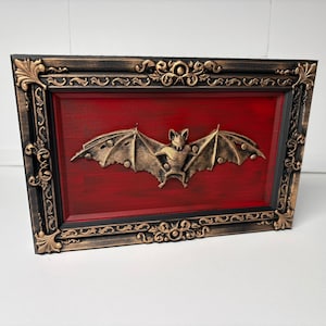 May include: A rectangular wall art piece featuring a central gold-colored bat relief mounted on a deep red background. The artwork is enclosed in a dark, ornate, distressed-finish frame with intricate scrollwork and leaf-like corner details.
