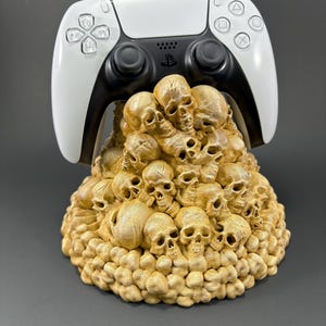 May include: A white PlayStation 5 controller sits on a skull-shaped stand. The stand is made of a light brown material and is covered in small skulls.