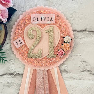 May include: A pink and gold glitter badge with the name "Olivia" and the number "21". The badge has a heart shape in the center and is decorated with small flowers and ribbons.