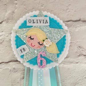 Pode incluir: Uma faixa de aniversário de feltro azul e branco com uma estrela de glitter prateada, o rosto de uma menina e o texto "Olivia faz 6 anos".