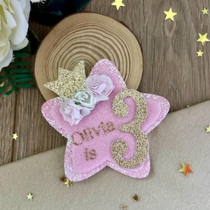 Könnte beinhalten: Rosa sternförmige Geburtstagsdekoration mit dem Namen "Olivia" und der Zahl "3" in Goldglitzer. Verziert mit einer goldenen Glitzerkrone und kleinen rosa und silbernen Blumen. Die Dekoration befindet sich auf einer Holzscheibe.
