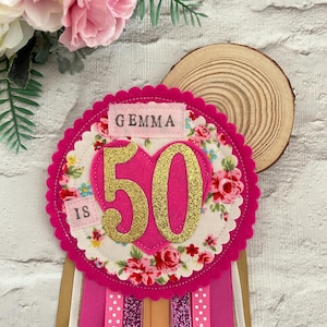 May include: A pink and floral 50th birthday badge with the name "Gemma". The badge features a heart shape with the number "50" in gold glitter, surrounded by a floral pattern and pink ribbons.