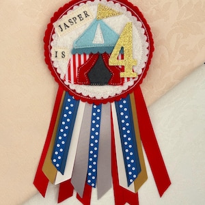 May include: A handmade felt birthday badge with a red scalloped edge. The badge features a circus tent, the number 4 in gold glitter, and the text "JASPER IS". Red, white, blue, and gold ribbons hang from the bottom.