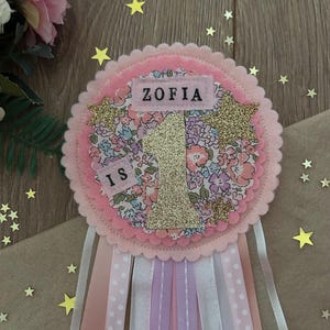 May include: A pink and floral fabric rosette with the name "ZOFIA" and the number "1" in gold glitter. The rosette has a scalloped edge and is adorned with ribbons in various shades of pink and white. Decorative stars are scattered around the rosette.