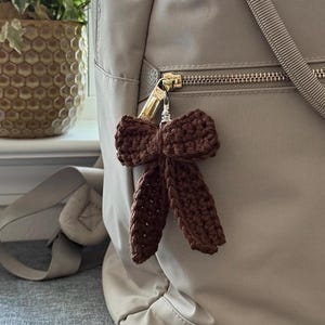May include: A brown crocheted bow keychain hanging from a beige handbag. The bow is made of a textured yarn and has two long, dangling loops. The handbag has a gold zipper and a beige strap.