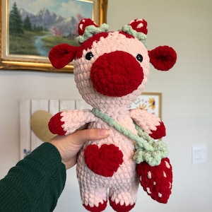 May include: A handmade crochet strawberry cow plushie. The cow is primarily pink with red accents, including a heart on its side, a strawberry nose, and strawberry-shaped ears. It carries a strawberry bag with a green strap.