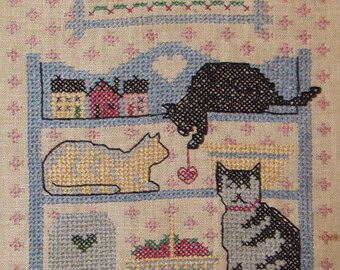 May include: Cross-stitch embroidery of a shelf with three cats and a basket of strawberries. The top shelf has a sign with the alphabet, the middle shelf has three cats, and the bottom shelf has a basket of strawberries. The background is a light beige fabric with a pink floral pattern.