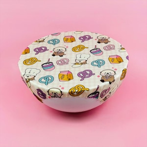 May include: A white bowl with a reusable food wrap featuring a pattern of brown otters, pretzels, and milk cartons. The otters are wearing chef hats and holding baking utensils.