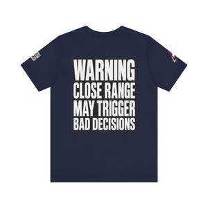 Puede incluir: Una camiseta azul marino de manga corta con texto blanco en la parte posterior que dice: WARNING CLOSE RANGE MAY TRIGGER BAD DECISIONS. La camiseta incluye un pequeño gráfico de código QR en la manga izquierda y un logo de estrella en la derecha.