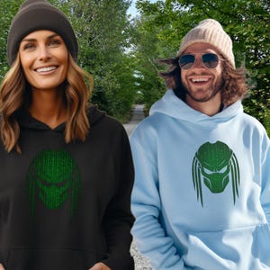 Peut inclure: Deux personnes portant des sweats à capuche assortis avec un motif Predator vert. L'un est noir, l'autre bleu clair. Les deux sourient. En arrière-plan, un chemin bordé d'arbres.