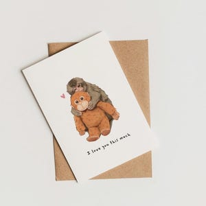 May include: A greeting card featuring an illustration of two monkeys hugging. One monkey is brown, the other is gray. The text "I love you this much" is printed below the illustration. The card is on a brown envelope.