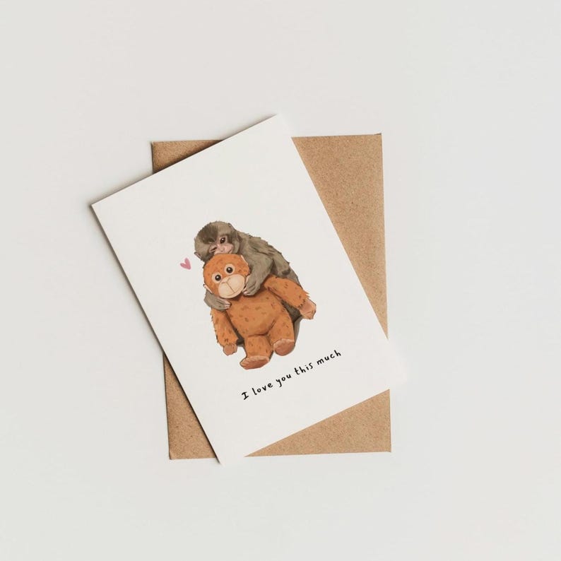 May include: A greeting card featuring an illustration of two monkeys hugging. One monkey is brown, the other is gray. The text "I love you this much" is printed below the illustration. The card is on a brown envelope.