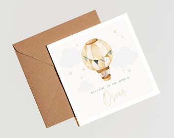 Puede incluir: Tarjeta de felicitación cuadrada con una ilustración de acuarela de un globo aerostático, nubes y estrellas. El texto "Welcome to the World" y el nombre "Oscar" están impresos en dorado. Un sobre marrón está detrás de la tarjeta.