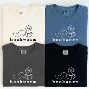 May include: Four folded t-shirts in cream, blue, charcoal, and black. Each shirt features a white graphic of a worm wearing glasses and reading a book, with the word "bookworm" below. The shirts are casual wear.