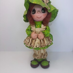 May include: A green and brown fabric doll with a green hat and brown hair. The doll is wearing a green dress with a brown apron and brown shoes.