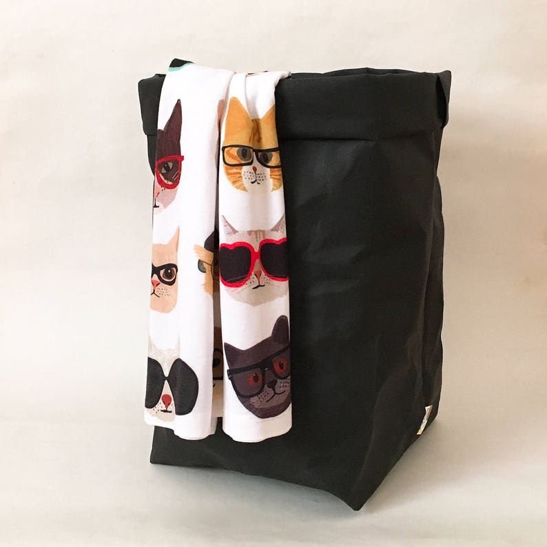 Puede incluir: Un cesto de ropa de tela negra con una bolsa de tela blanca con un patr&oacute;n de gatos que llevan gafas de sol y gafas. La bolsa tiene un fondo blanco con una variedad de caras de gatos con gafas de sol y gafas de diferentes colores.