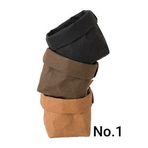 Puede incluir: Tres bolsas de papel, una negra, una marr&oacute;n y una beige, apiladas una encima de la otra. Las bolsas est&aacute;n todas dobladas por la mitad y tienen un fondo redondeado. El texto "No. 1" es visible en la esquina inferior derecha de la imagen.
