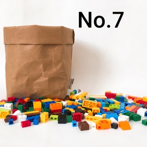 Puede incluir: Una bolsa de papel marr&oacute;n con un mont&oacute;n de bloques de construcci&oacute;n de pl&aacute;stico de colores delante. La bolsa est&aacute; arrugada y tiene una etiqueta en el lateral. Los bloques son de varios colores, incluyendo rojo, azul, verde, amarillo y naranja. El texto "No. 7" est&aacute; escrito en negro sobre un fondo blanco encima de la bolsa.