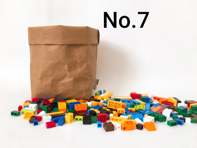 Puede incluir: Una bolsa de papel marr&oacute;n con un mont&oacute;n de bloques de construcci&oacute;n de pl&aacute;stico de colores delante. La bolsa est&aacute; arrugada y tiene una etiqueta en el lateral. Los bloques son de varios colores, incluyendo rojo, azul, verde, amarillo y naranja. El texto "No. 7" est&aacute; escrito en negro sobre un fondo blanco encima de la bolsa.
