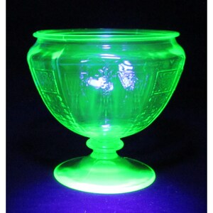 May include: A vibrant, glowing neon green glass bowl with a pedestal base. The bowl features a wide, circular rim and a textured, patterned exterior surface that emits a bright, luminous light, characteristic of uranium glass under UV lighting.