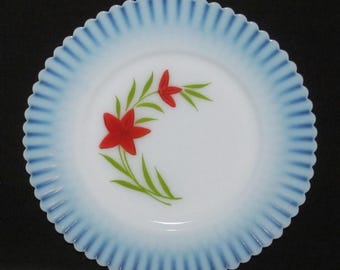 May include: White plate with a blue scalloped rim and a floral design. The plate features a white center with red flowers and green leaves. The rim has a ribbed texture.