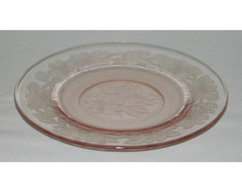 May include: A pink glass dessert plate with a floral design. The plate has a raised rim and a center design of a flower with leaves.