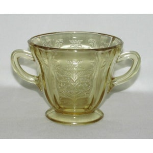 May include: A yellow glass sugar bowl with two handles and a pedestal base. The bowl features an embossed floral design on the exterior. The glass has a slightly translucent quality.