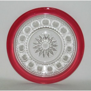 May include: A clear glass plate with a starburst design in the center and a red rim. The plate has a decorative pattern of small circles around the edge.