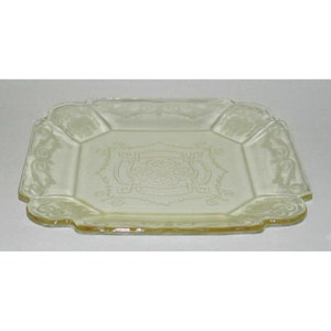 May include: A square, yellow glass serving plate with an intricate, etched design. The plate has a scalloped edge and a raised center.