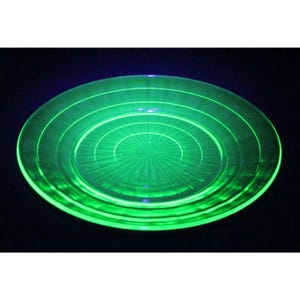 May include: A glowing green glass plate with concentric circles and radial lines emanating from the center. The plate has a circular shape and is illuminated, creating a vibrant, eye-catching effect. The plate is likely vintage or antique.