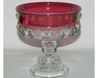 May include: A clear glass pedestal with a ruby red bowl. The pedestal has a textured design and the bowl has a smooth, shiny finish.