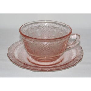 May include: A pink glass teacup and saucer set with a delicate floral pattern. The cup and saucer are both made of clear glass with a pink tint. The pattern is embossed on the glass and features a repeating design of flowers and leaves.