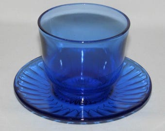 May include: A cobalt blue glass teacup and saucer set. The teacup is cylindrical with a smooth rim, and the saucer has a ridged design. The set is made of transparent glass, and the color is consistent throughout.
