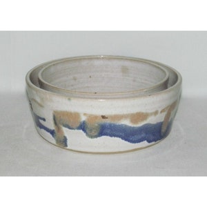 May include: A set of two ceramic bowls, one nested inside the other. The larger bowl has a white glaze with blue and brown abstract designs. The smaller bowl is plain white.