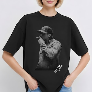 Peut inclure: T-shirt noir avec une image en noir et blanc d'une personne fumant une cigarette, portant une casquette de baseball et une chemise boutonnée. L'image comporte une signature en bas à droite.