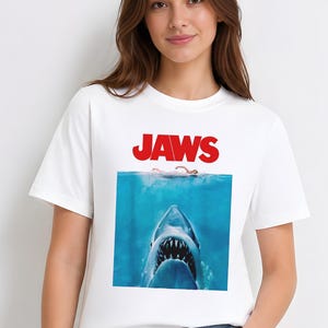 Puede incluir: Camiseta blanca con el cartel de la película "Tiburón". El cartel muestra la boca abierta de un tiburón bajo el agua, con un nadador encima. La palabra "JAWS" está impresa en rojo en la parte superior.