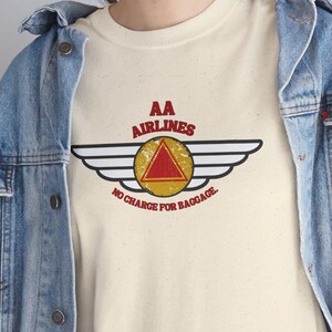 May include: Cream-colored t-shirt featuring a vintage-style airline logo with the text "AA AIRLINES" above a triangle and wings design. Below the logo, the text reads "NO CHARGE FOR BAGGAGE". The shirt is worn with a denim jacket.
