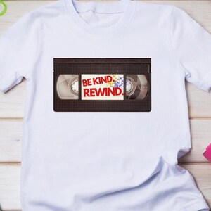 May include: White t-shirt featuring a VHS tape graphic with the text "BE KIND REWIND." The shirt is on a light wood surface with blue jeans, party favors, and colourful straws. A casual, fun tee.