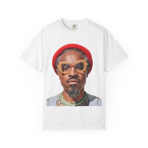 Op de afbeelding: Wit T-shirt met een portret van een man met een rode muts, een grote beige bril en een sik. De man draagt ook een gestreept shirt en een groene ketting. Het T-shirt heeft een klein label op de bovenrug.