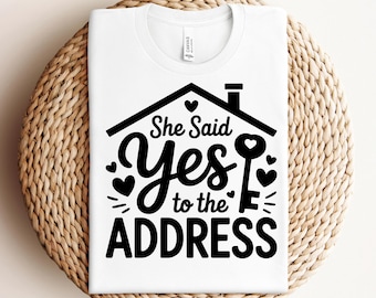 Puede incluir: Camiseta blanca con un diseño negro que dice "She Said Yes to the Address". El diseño incluye una casa, corazones y una llave. La camiseta se muestra sobre una superficie tejida de color marrón claro.